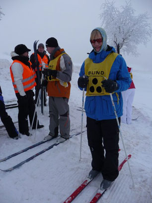 Finn wartet mit angeschnallten Skiern und den Stöcken in den Händen auf den Start. Im Hintergrund ist Max mit Frau Cjaika auf Skiern zu sehen.