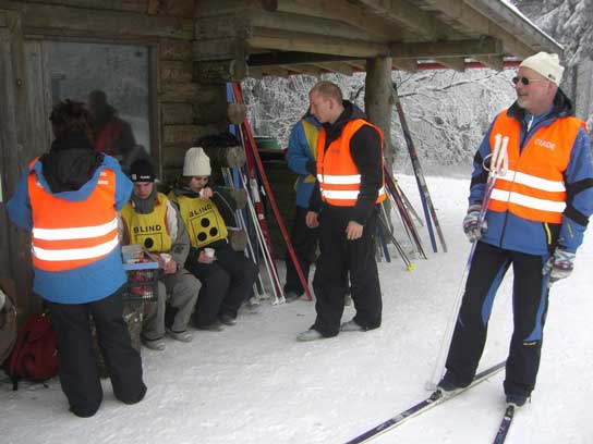 An einer Hütte am Skistadion Winterberg sitzen Max und Sinem und machen Pause. Frau Hasbargen, Holger und Günter stehen dabei.