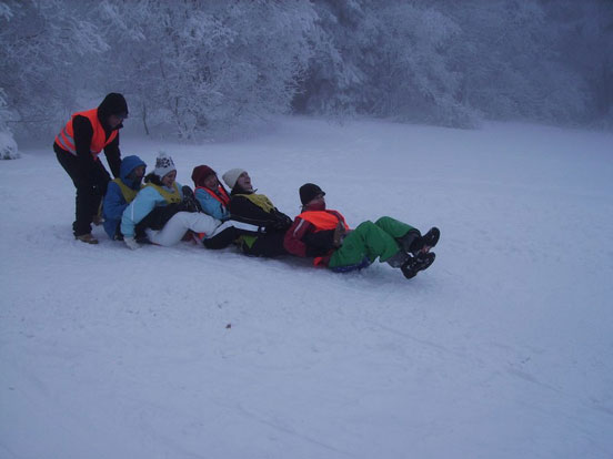 Finn, Vivien, Sinem, Frau Thoma und Lisa sitzen hintereinander an einem Hang im Schnee, um so zu rodeln. Christoph schiebt sie von hinten an.