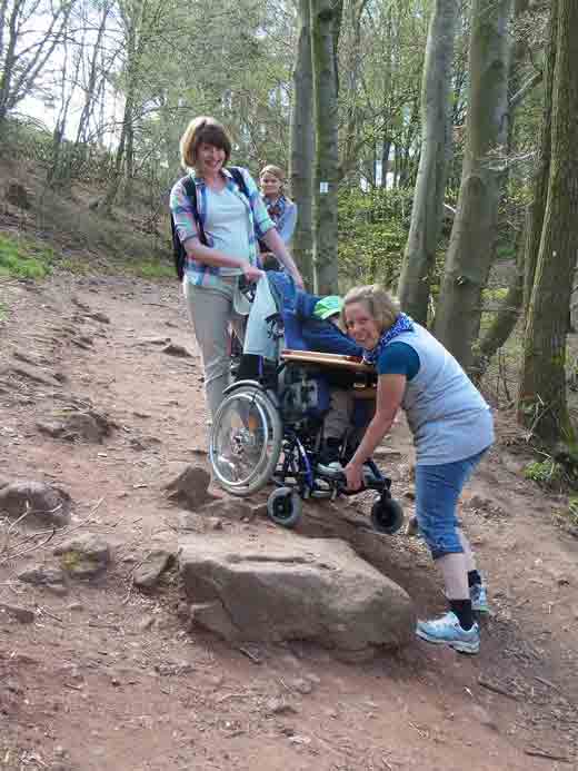Frau Kähler und Frau Nettersheim tragen Fabian in seinem Rollstuhl über einen dicken Stein, der im Weg liegt.