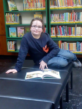 Angelina sitzt mit einem Buch auf einer Bank und bereitet sich auf den Lesewettbewerb vor. 