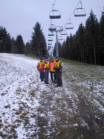 Matthias, Andreas, Mustafa und Günter stehen mitten auf einer Piste mit wenig Schnee an einem Sessellift.