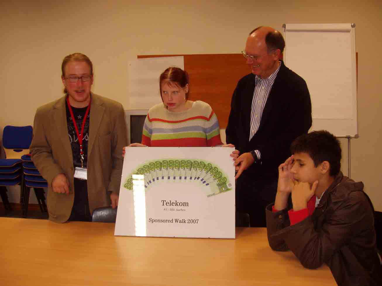 Dennis Ürlings übergibt 18 Scheine im Wert von je 100  an die Schülersprecherin Angela Quentmeier. Die Scheine sind im Halbkreis auf einem Bild angeordnet. Herr Franz schaut lächelnd zu.